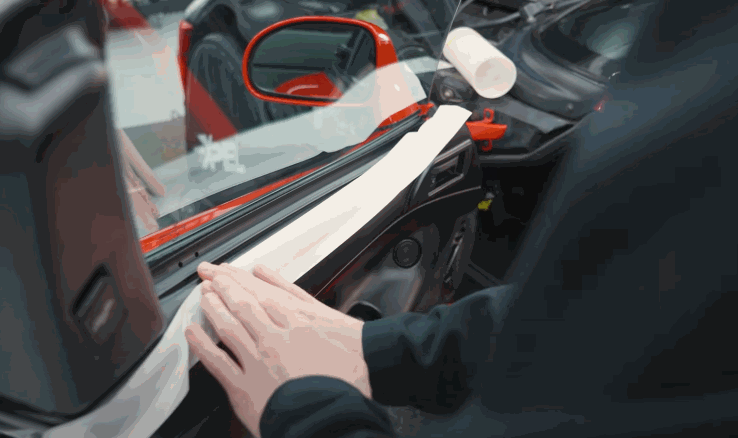 Technician carefully aligns and presses window tint film onto the inside of a car door window near a red side mirror. The film is partially peeled back showing the installation process of window tint for a clean finish.