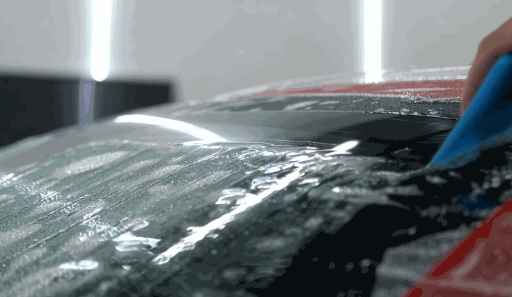 Close view of a tool smoothing liquid across a car windshield as excess moisture is pushed away during installation. Reflections of overhead lights highlight the wet surface and even pressure being applied. This step ensures a clean and properly adhered windshield finish.