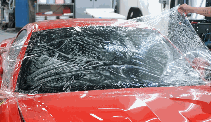 Red car windshield covered in soapy solution as a clear film is carefully laid across the surface by hand. The film stretches over the curved glass while trapped water and air are visible underneath. This shows the early stage of windshield film application.