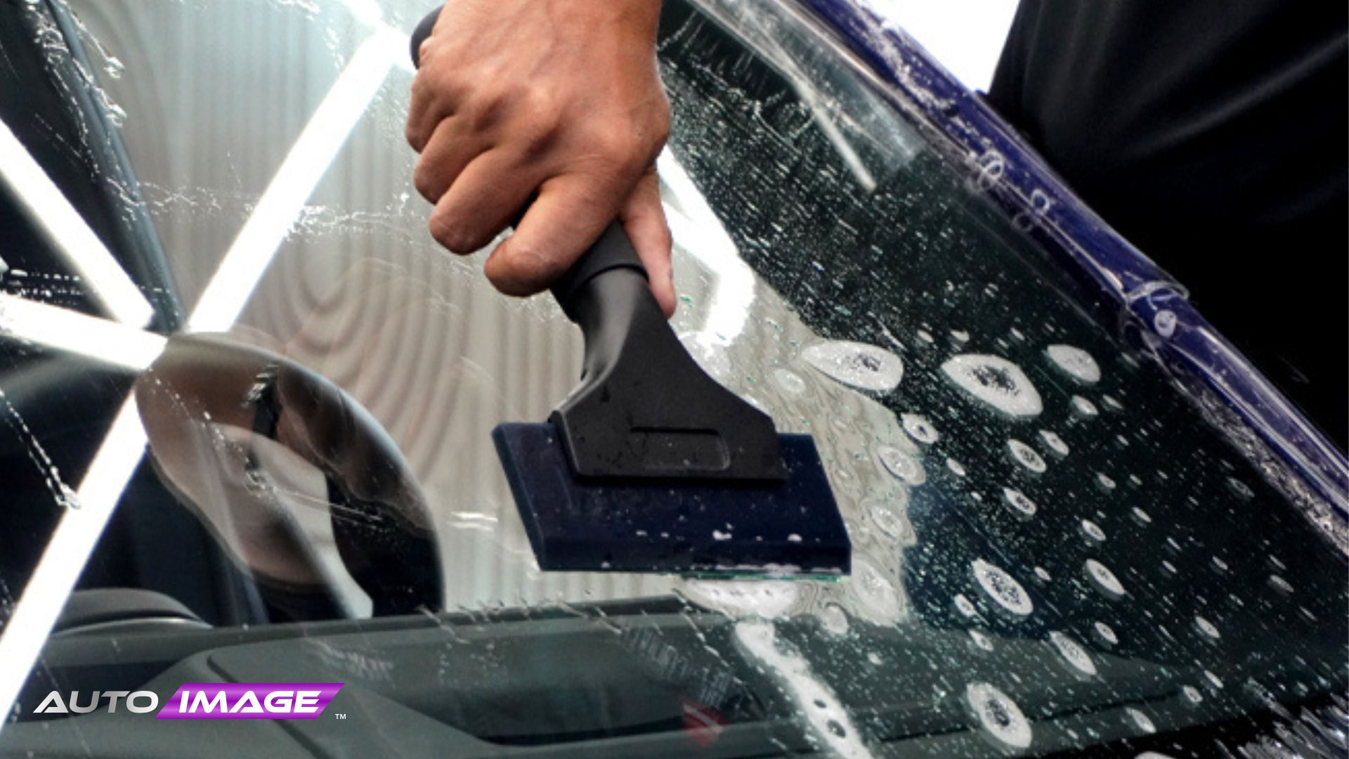 Close up of a hand using a squeegee to remove soapy water from a car windshield during tint or film installation. Bright shop lights reflect across the glass while bubbles are pushed out for a smooth finish. The process shows precision work on the windshield surface.