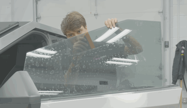 A worker holds up a sheet of ceramic window tint against a car window, with soapy water droplets visible across the glass surface. The partially installed film shows how the tint will reduce glare and improve privacy once secured.