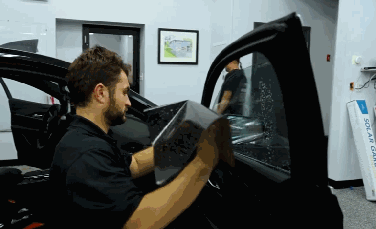 A technician installs ceramic window tint on a car door inside a workshop, carefully pressing the film onto the glass with both hands. Water droplets and reflections on the window show the tint being applied smoothly for a clean finish.