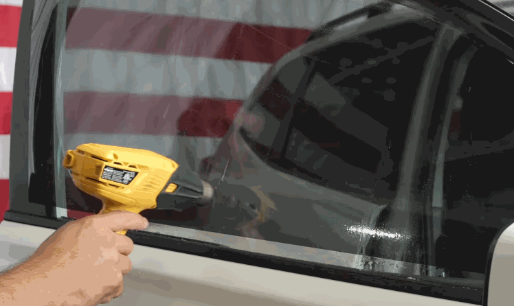 Yellow heat gun warming window tint film on a car side window to help it shrink smoothly against the glass.