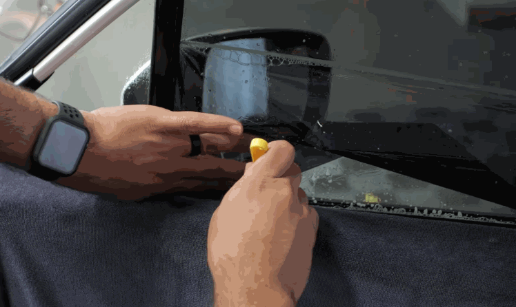 Hands using a yellow tool to tuck window tint film along the lower edge of a car side window.