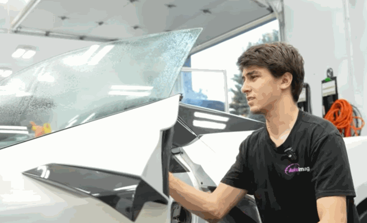 A technician peels old window tint from the rear window of a white car during window tint replacement. The film lifts away from the glass inside a bright auto shop.